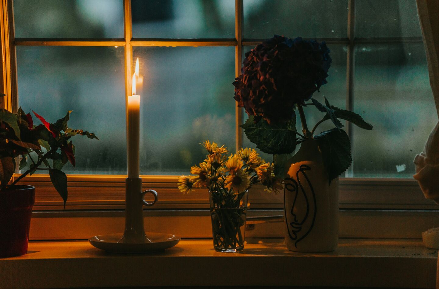 Lumânare aprinsă lângă o vază cu flori, lumină caldă de fereastră